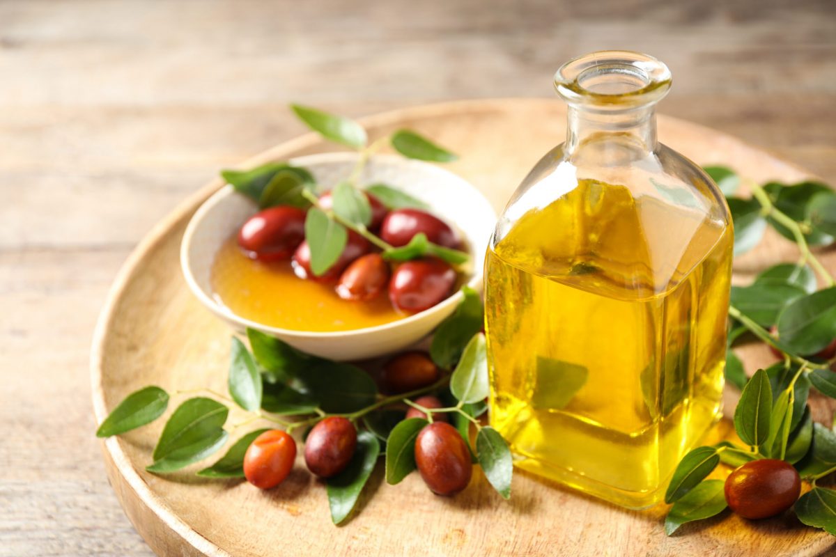 Flacon d'huile de jojoba en premier plan avec un petit bol contenant les fruits de jojoba en arrière plan