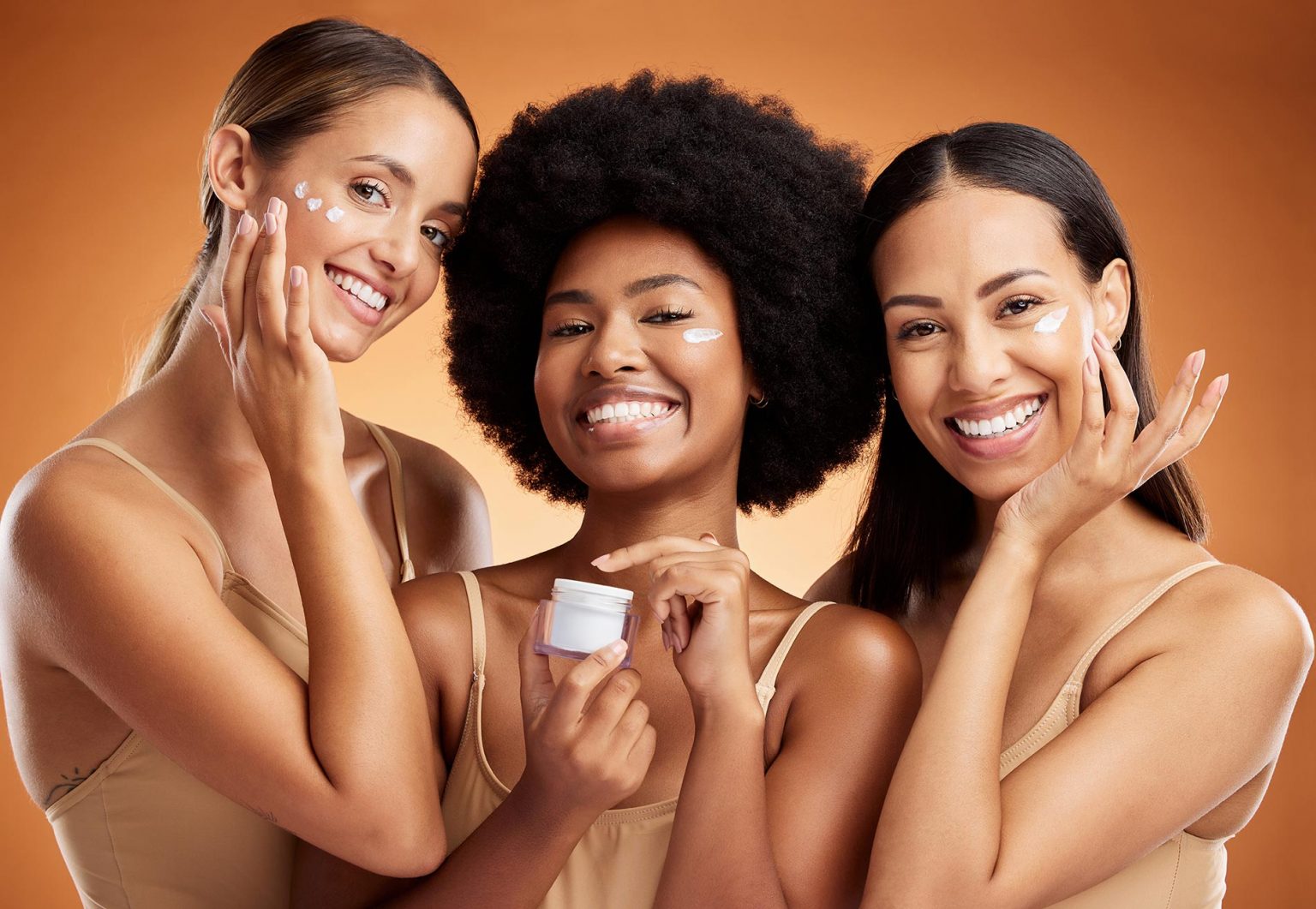 Plan buste de 3 femmes souriantes, de différentes ethnies, qui s'appliquent de la crème sur le visage